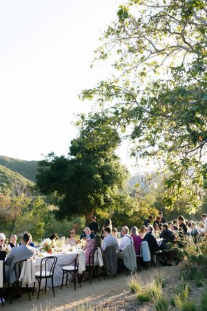 Long outdoor wedding reception table set among trees and mountains at sunset.