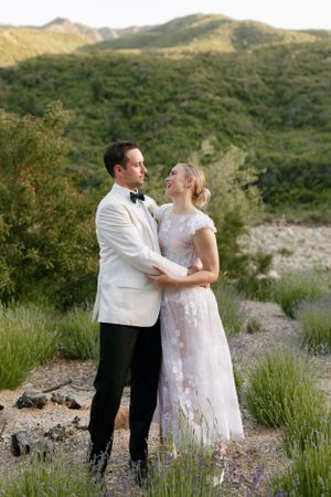 Couple in white formal wear embracing against mountainous backdrop.