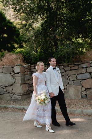 A couple in wedding attire walks together along a stone wall path.