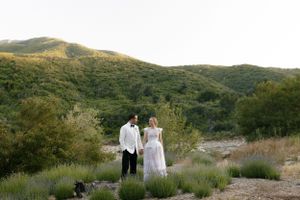 A couple in wedding attire stand together on a hillside with mountain landscape in background.