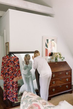 Two figures in white and blue outfits stand at an antique wooden dresser with a floral dress hanging nearby.