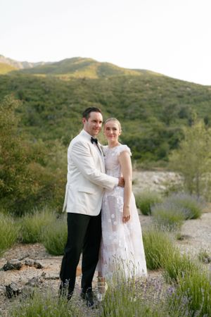 A couple in wedding attire pose together against a scenic mountain backdrop at sunset.