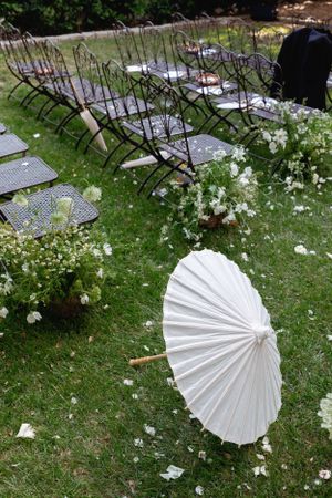 Metal folding chairs and a white paper parasol arranged for an outdoor wedding with scattered flower petals.