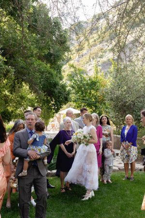 Wedding guests gather on a lush green lawn surrounded by trees during an outdoor celebration.