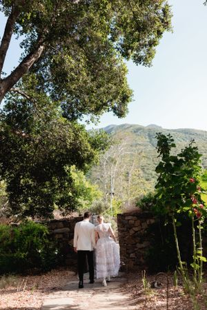 A couple walks along a scenic path with mountains and trees in the background.