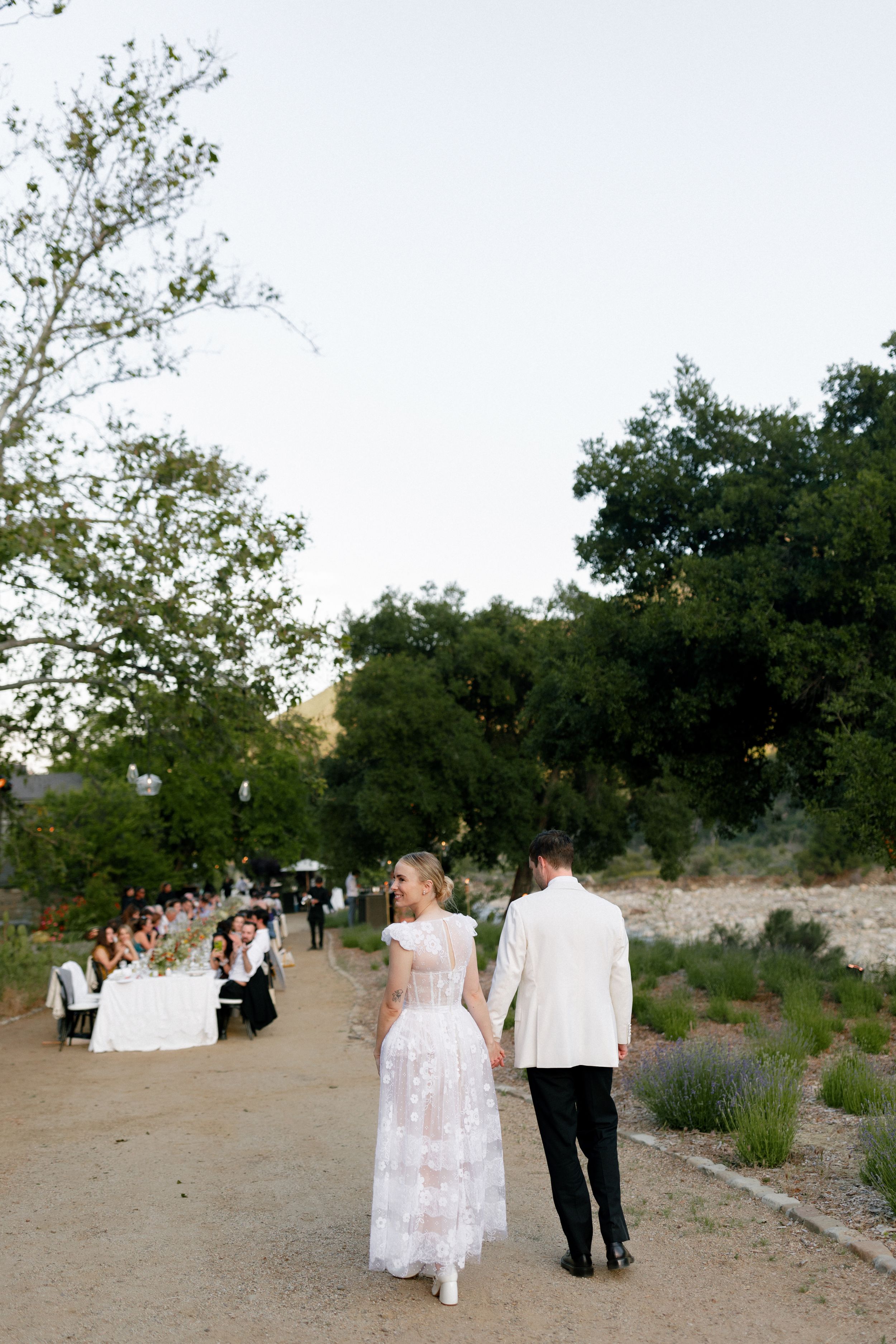 Newlyweds walk down a path as wedding guests dine at a long table in the distance.