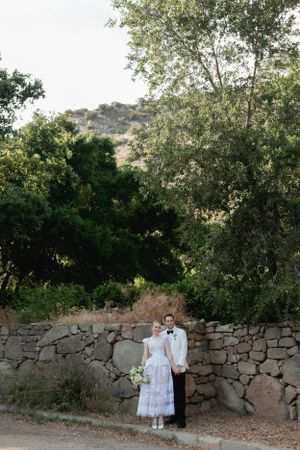 A couple poses against a rustic stone wall with trees and hills in the background.
