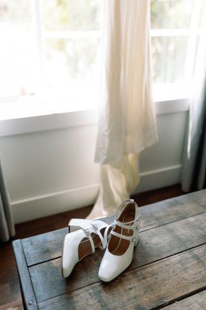 White vintage-style wedding shoes displayed on a rustic wooden surface near a window.