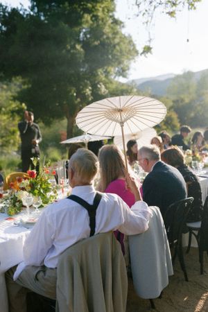 Outdoor wedding reception table with guests and a decorative white parasol in a scenic mountain setting.