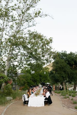 Long dinner table set up on garden path beneath overhanging trees at dusk.