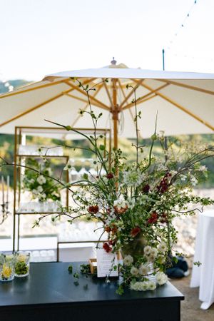 Large floral arrangement with white and burgundy flowers under cream-colored patio umbrella.