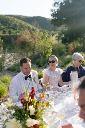 Outdoor wedding reception with guests dining at a beautifully decorated table surrounded by hills and greenery.