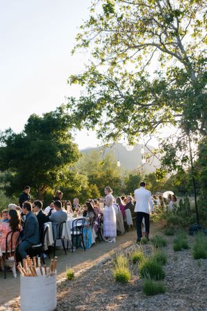 Outdoor wedding ceremony with guests gathered under trees at sunset in a rustic mountain setting.
