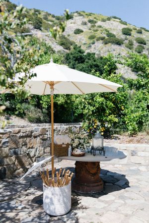 White market umbrella and vintage barrel setup against stone wall and mountain landscape.