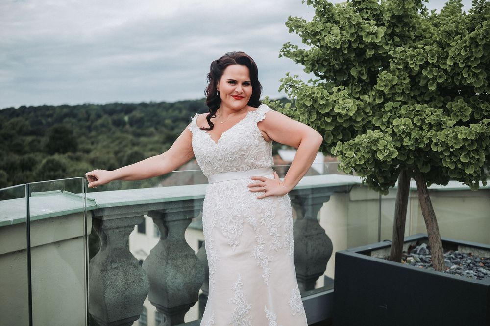 A bride in a white lace wedding dress poses on a balcony overlooking a scenic mountain landscape.
