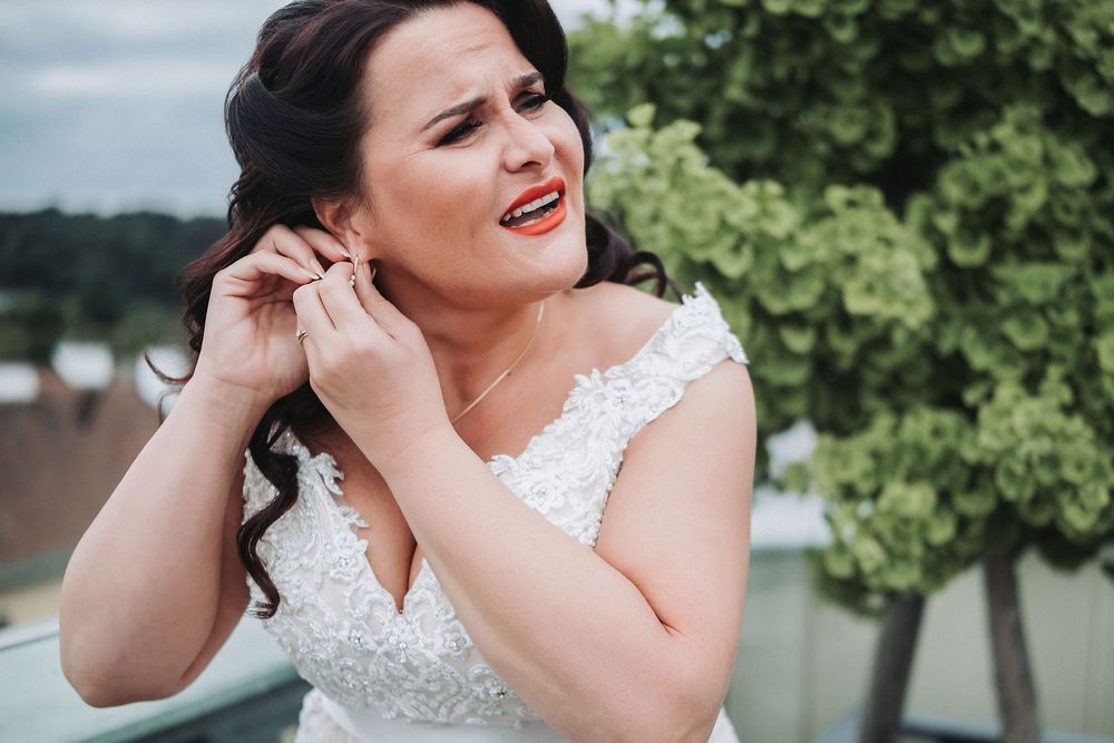 Bride in white lace dress poses outdoors with greenery and trees in the background.