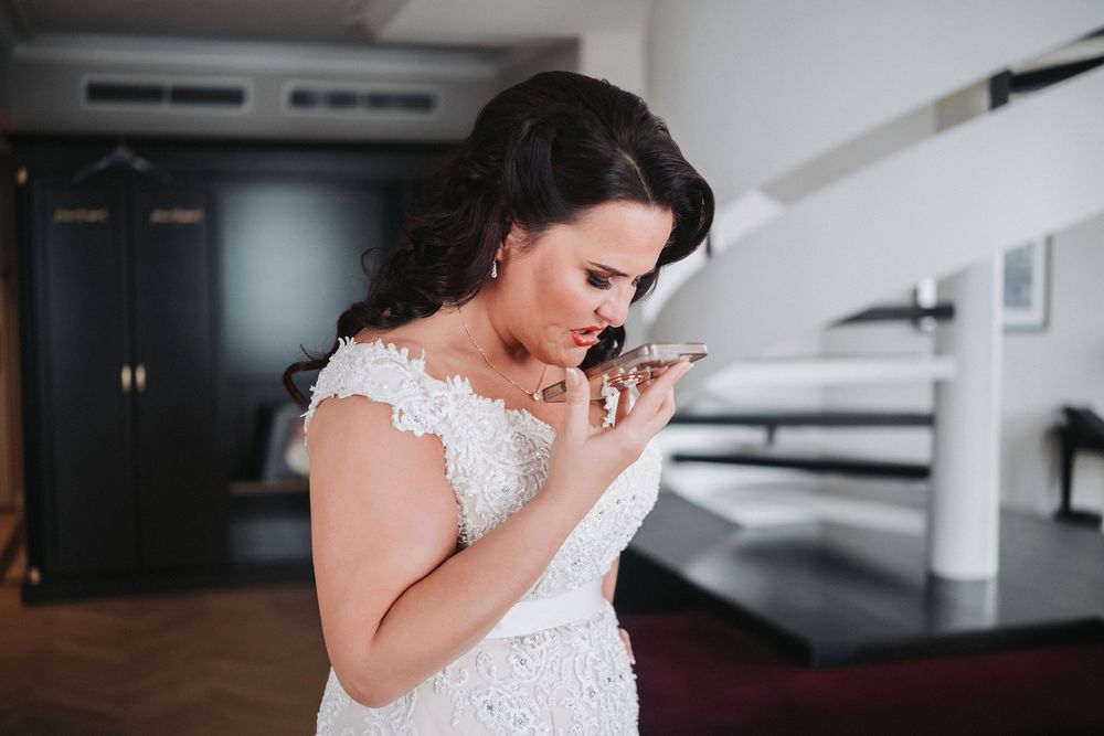A bride in a white lace wedding dress looks down while holding a bouquet in an elegant indoor setting.