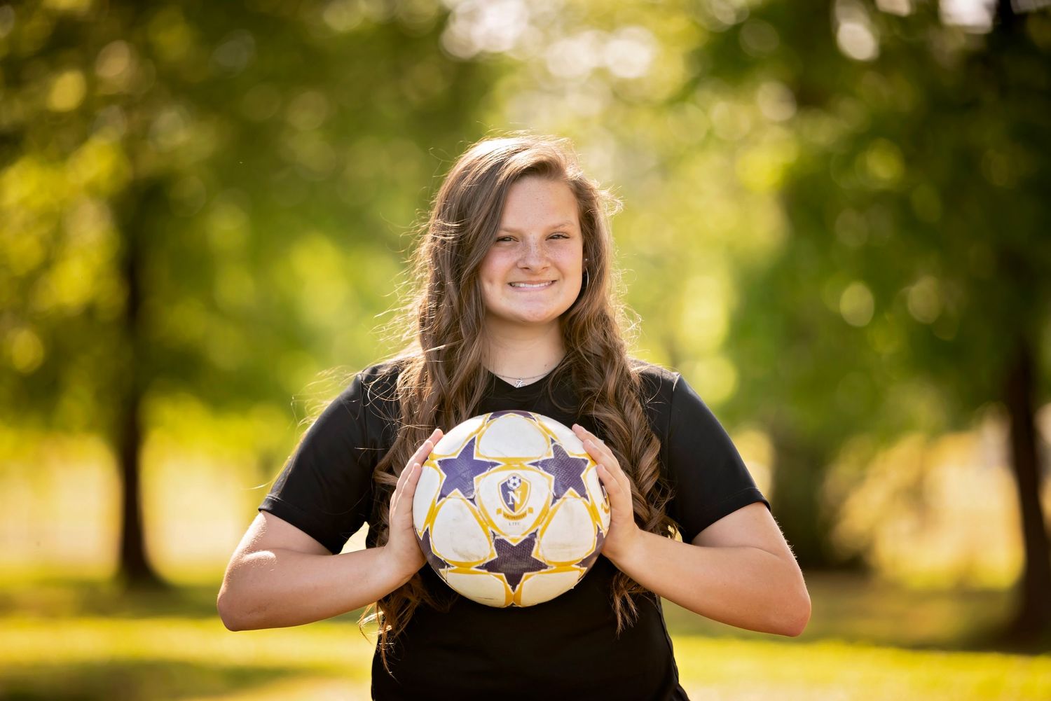 A soccer player holds a ball in front of them while standing in a sunlit park with trees in the background.