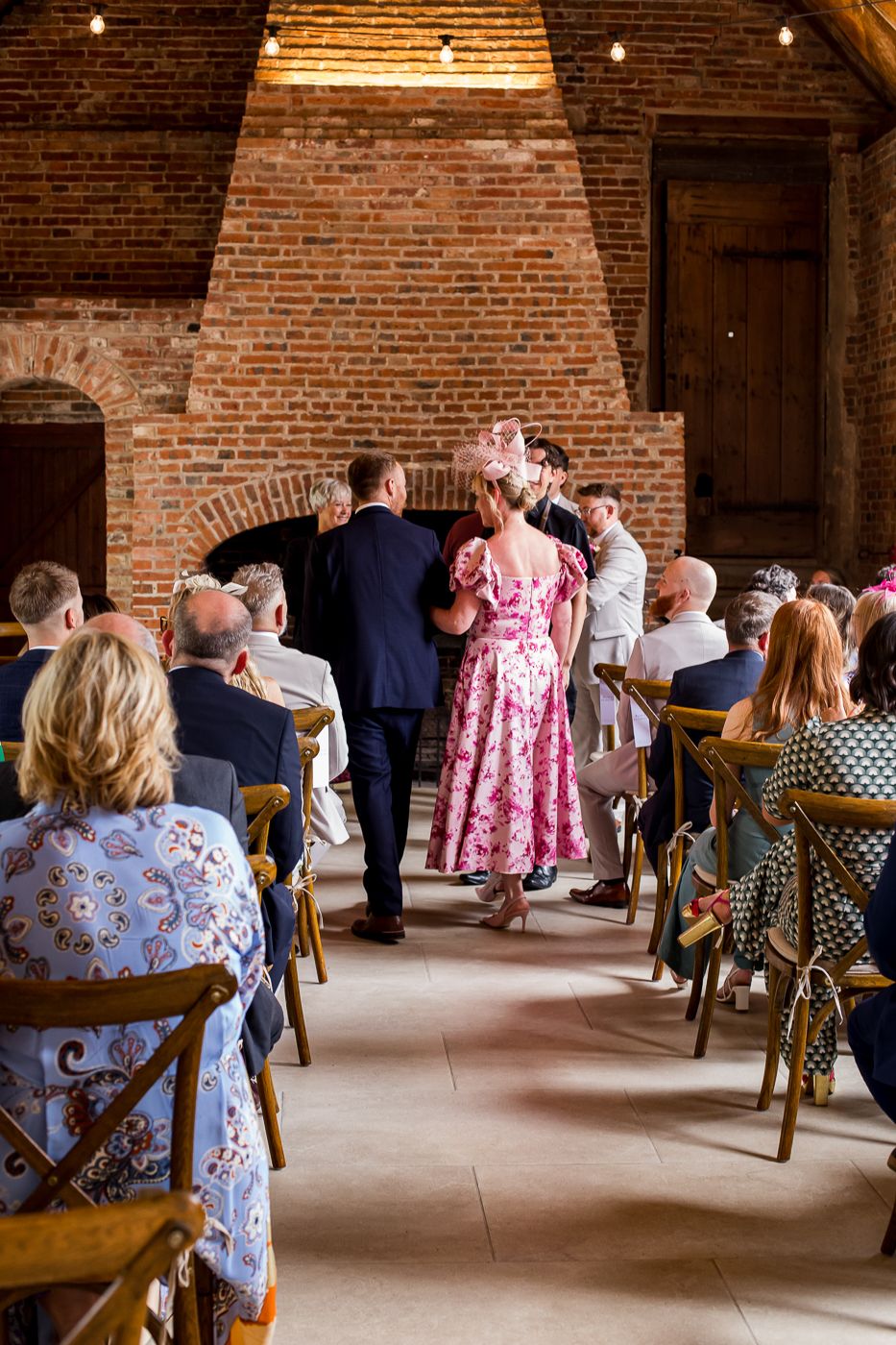 Wedding guests seated in wooden chairs watch a ceremony in a rustic brick venue.