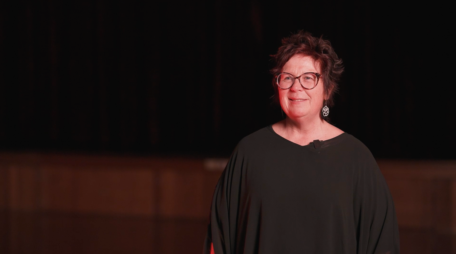 A performer in a black dress and red glasses stands on a dark stage.