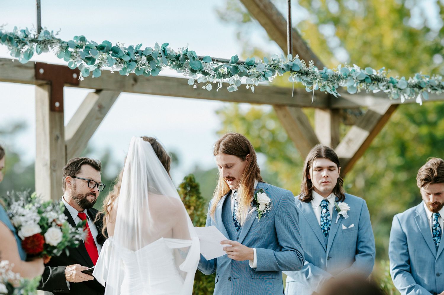 A wedding party celebrates outdoors with bridesmaids in burgundy dresses and groomsmen in gray suits.