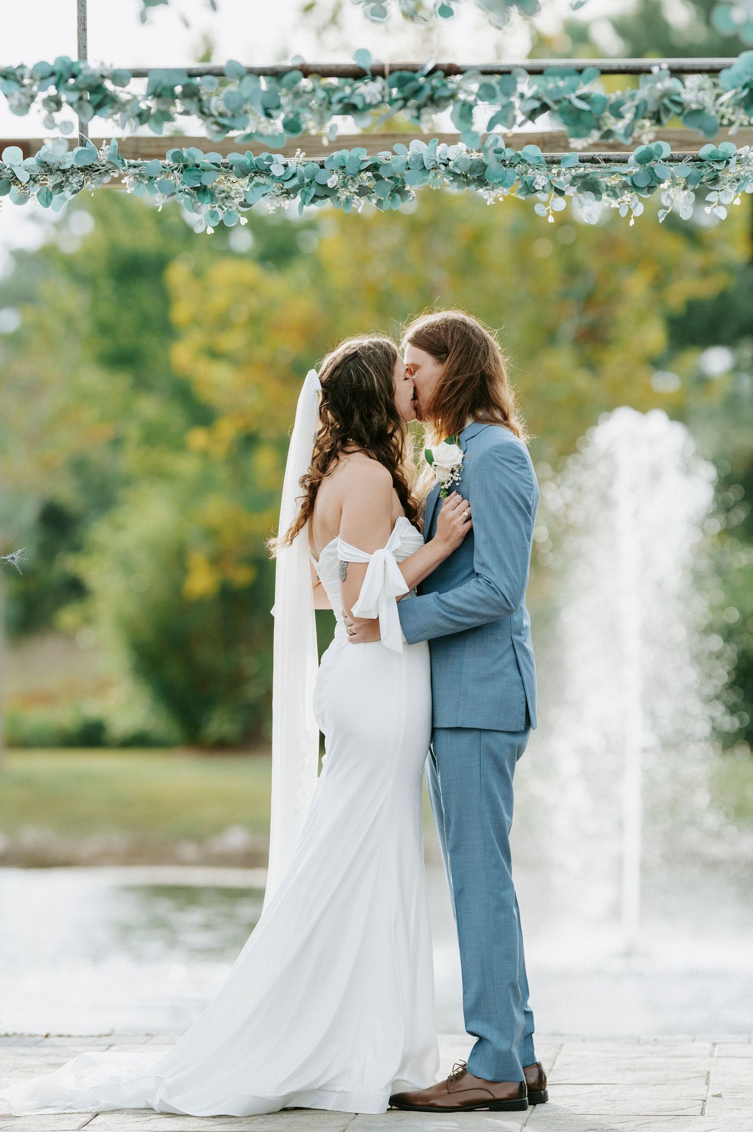 A romantic wedding couple shares a tender moment by a fountain in a garden setting with autumn trees in the background.