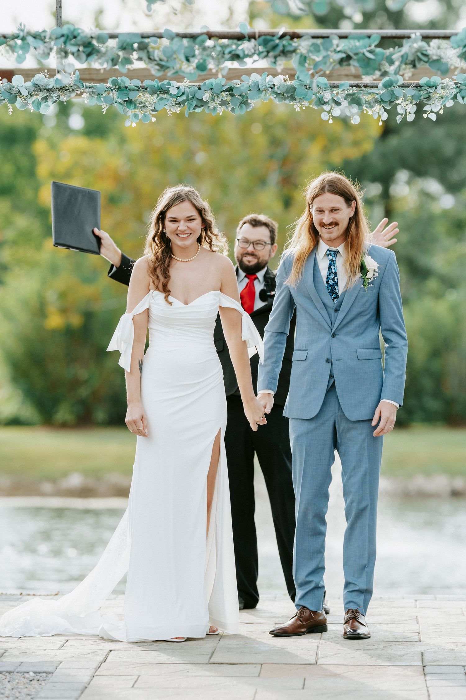 A wedding party walks together on a stone path near a lake during a sunny outdoor celebration.