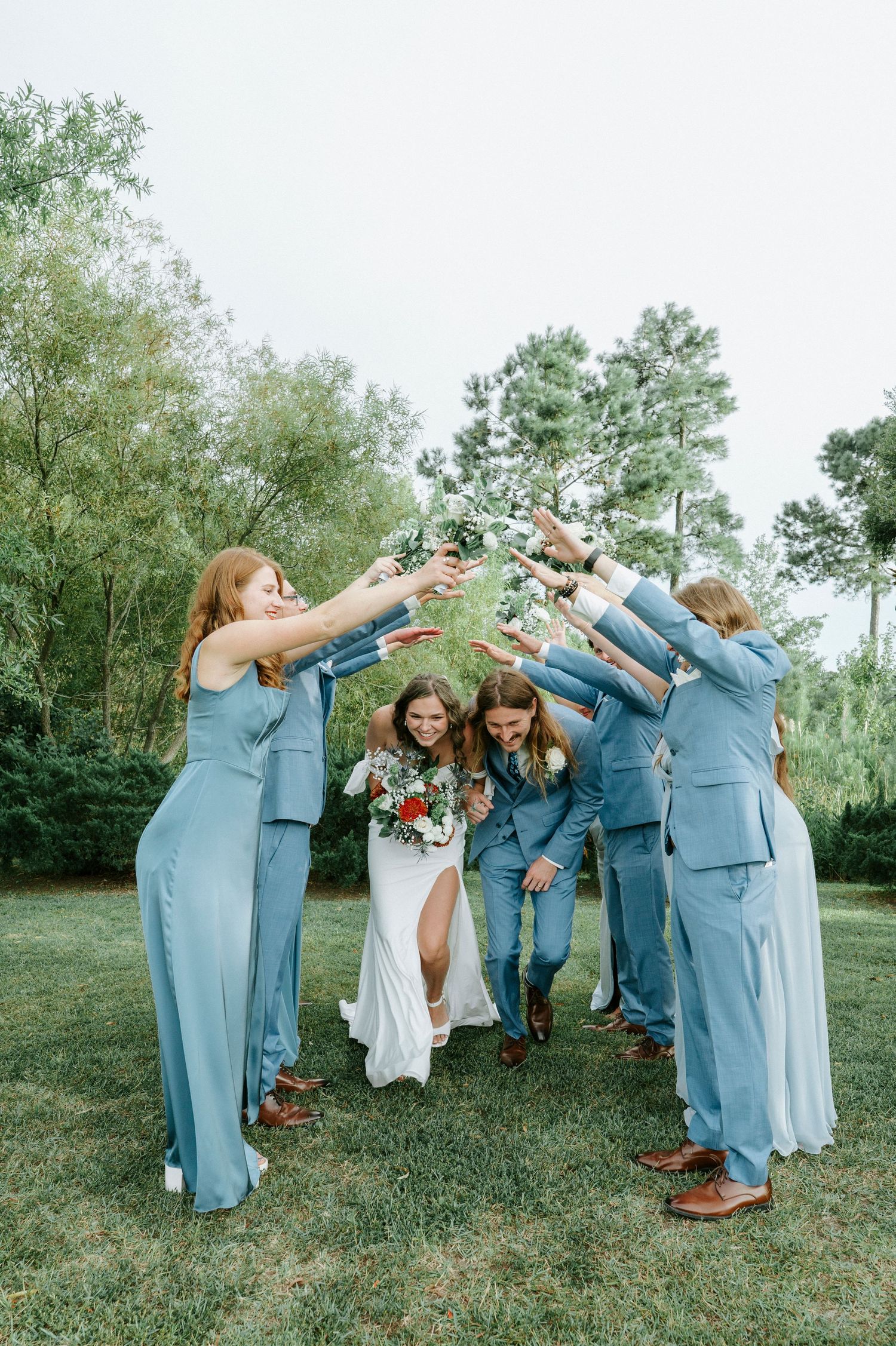 Wedding party wearing light blue outfits performs synchronized dance move outdoors in grassy field.