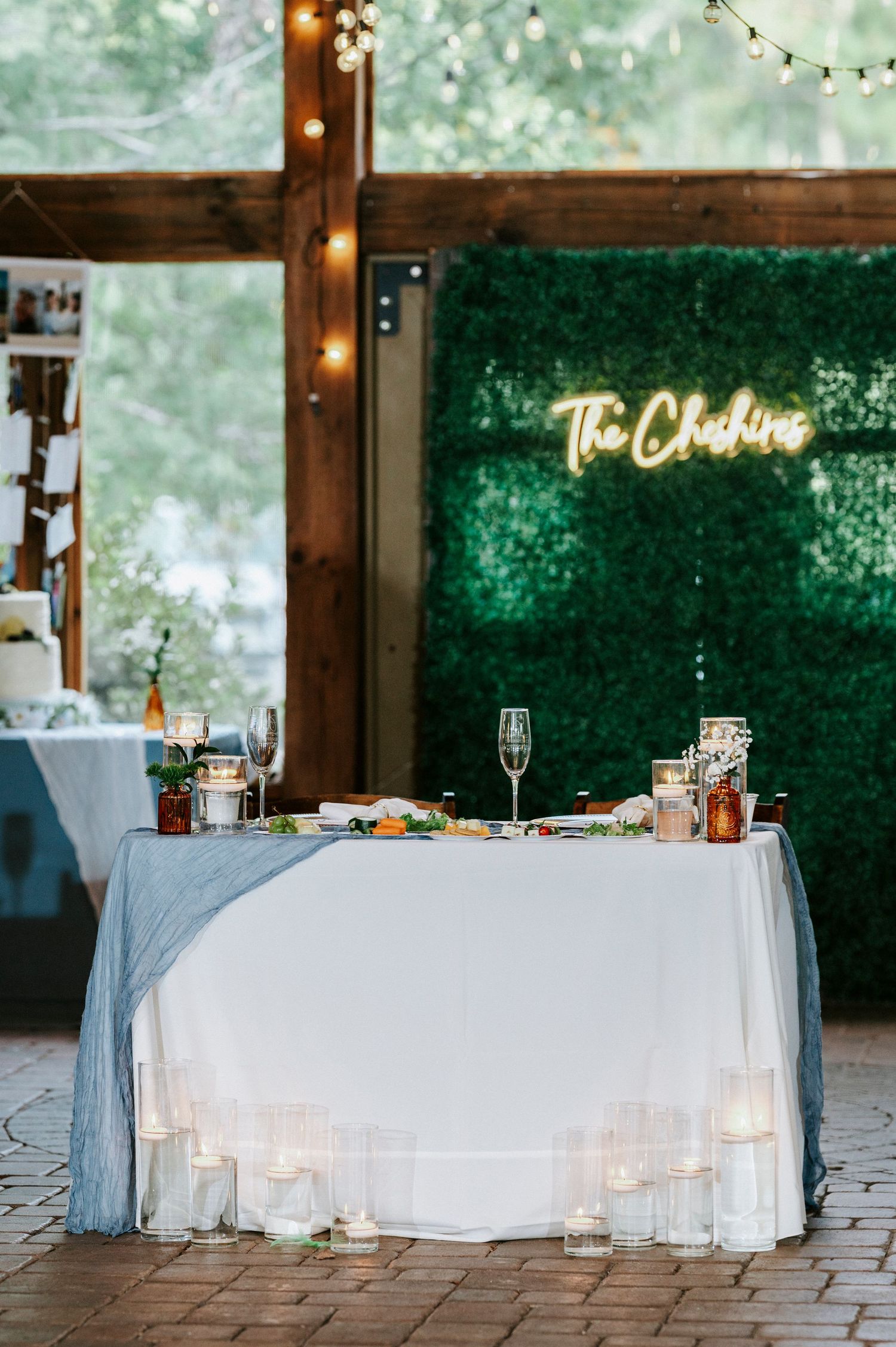 A wedding sweetheart table with white linens and string lights under a green neon sign in a rustic wooden venue.