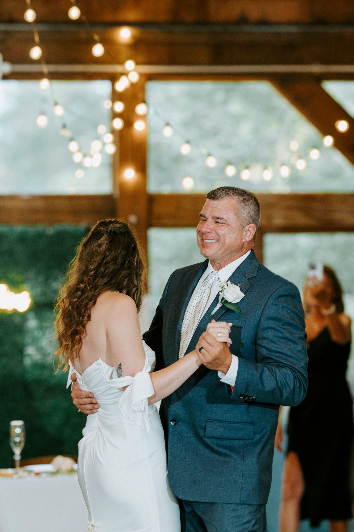 A couple shares a dance at a rustic barn wedding reception with twinkling string lights overhead.