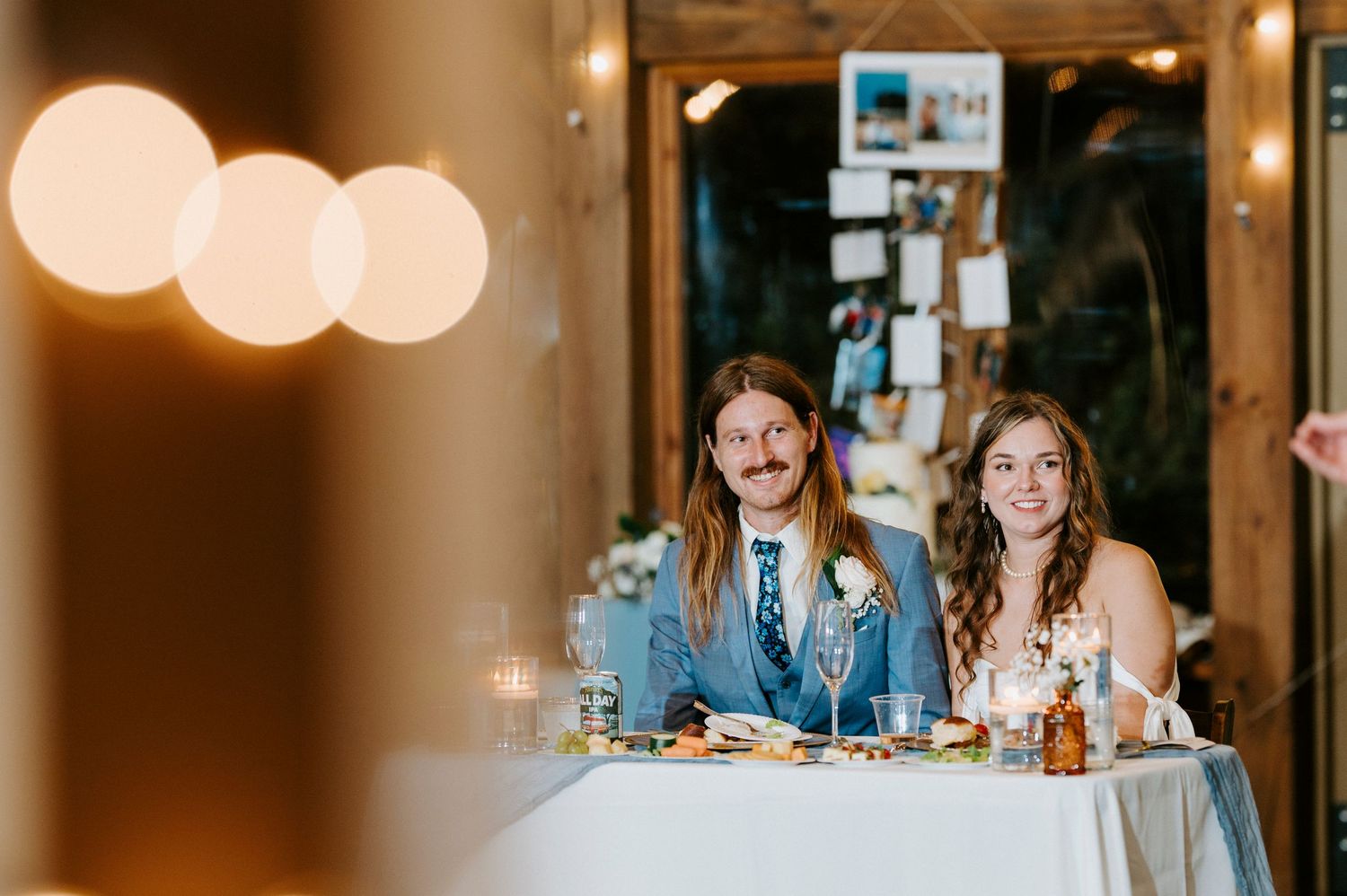 Wedding guests seated at a long dinner table with glowing pendant lights overhead during an evening reception.