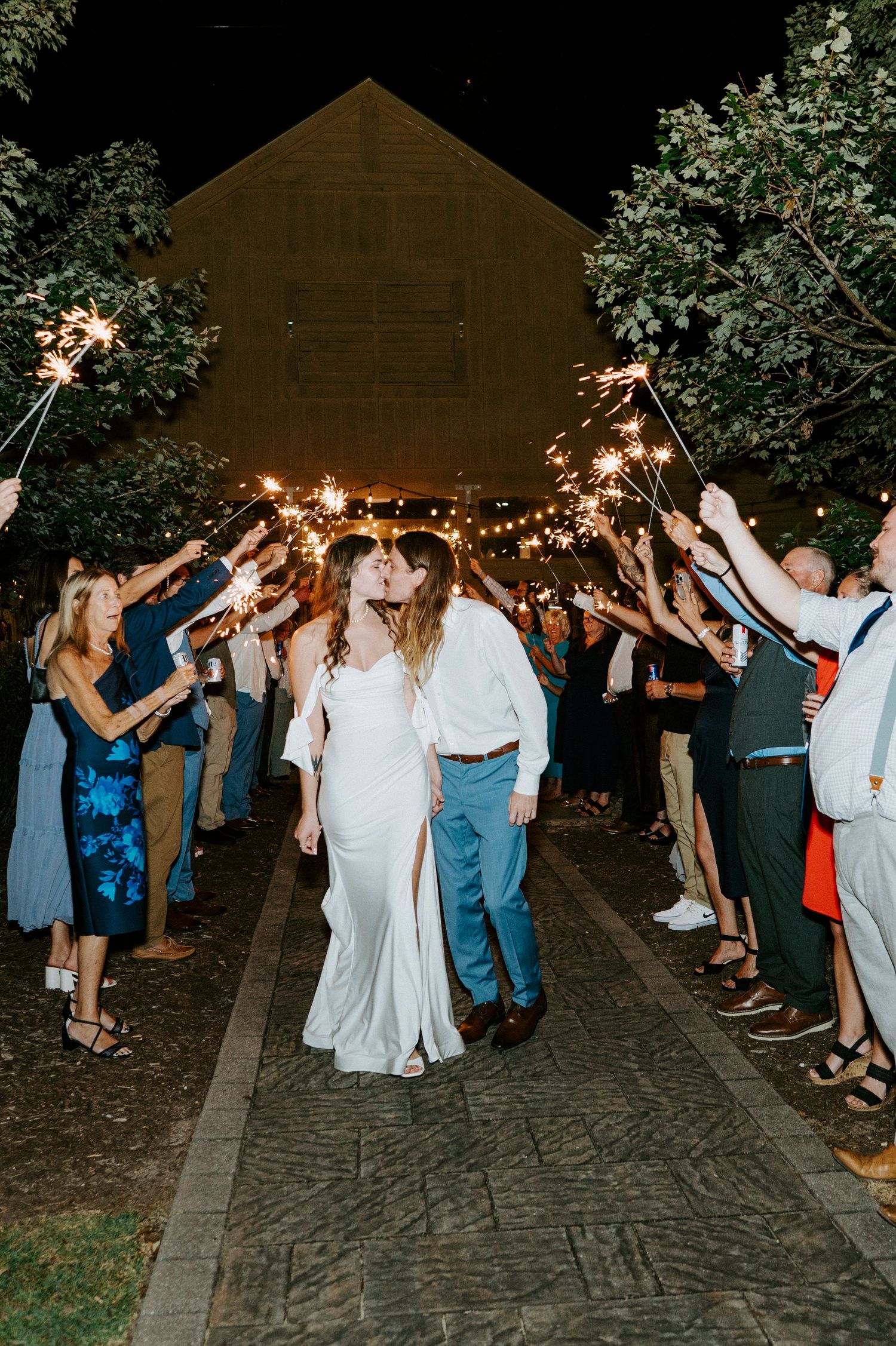 Wedding guests hold sparklers creating a magical nighttime send-off tunnel for the newlyweds on a cobblestone path.