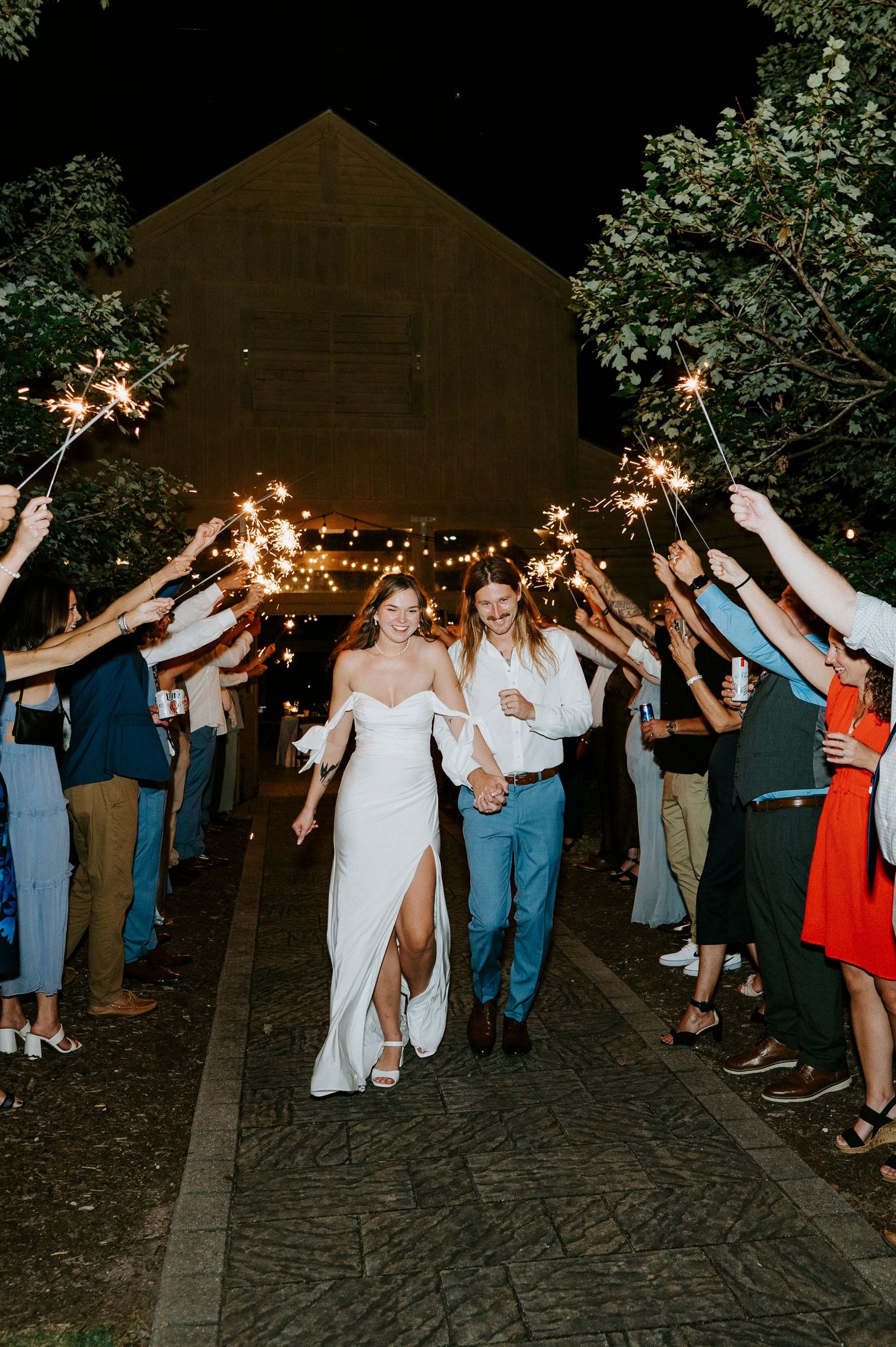 Wedding guests and couple form a celebratory exit tunnel on a cobblestone path at night under string lights.