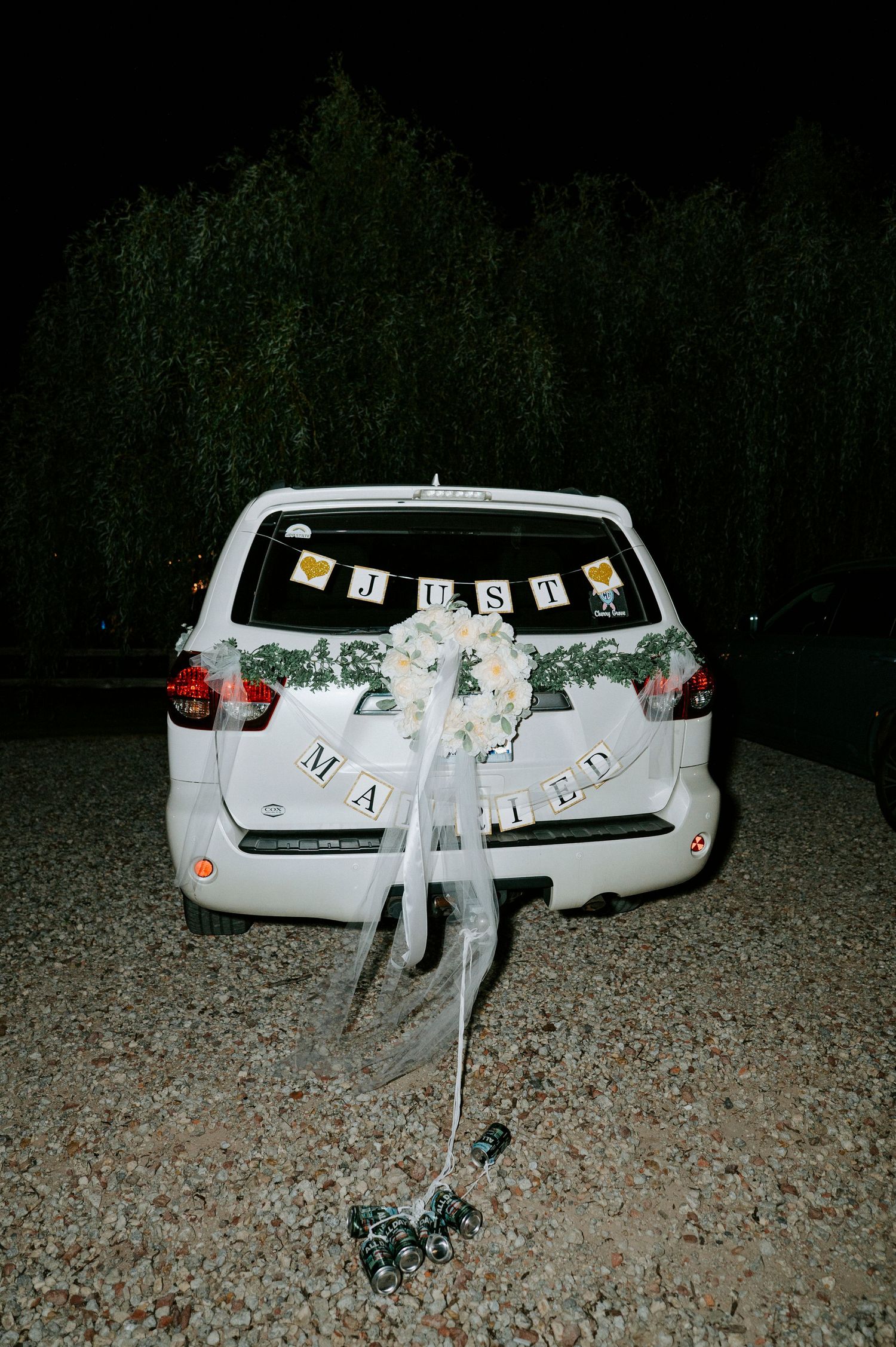 A white car decorated with wedding ribbons and 'Just Married' text parked on gravel at night.