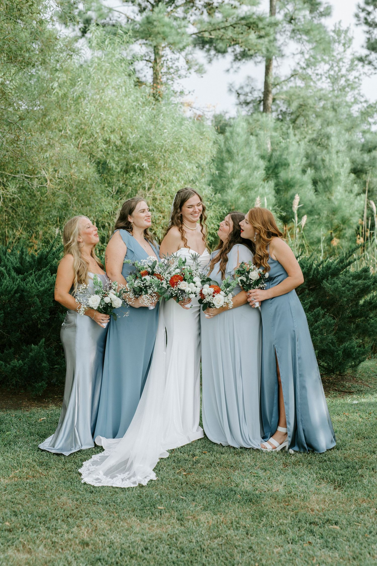 Bridesmaids in dusty blue dresses laugh together while holding bouquets in an outdoor garden setting.