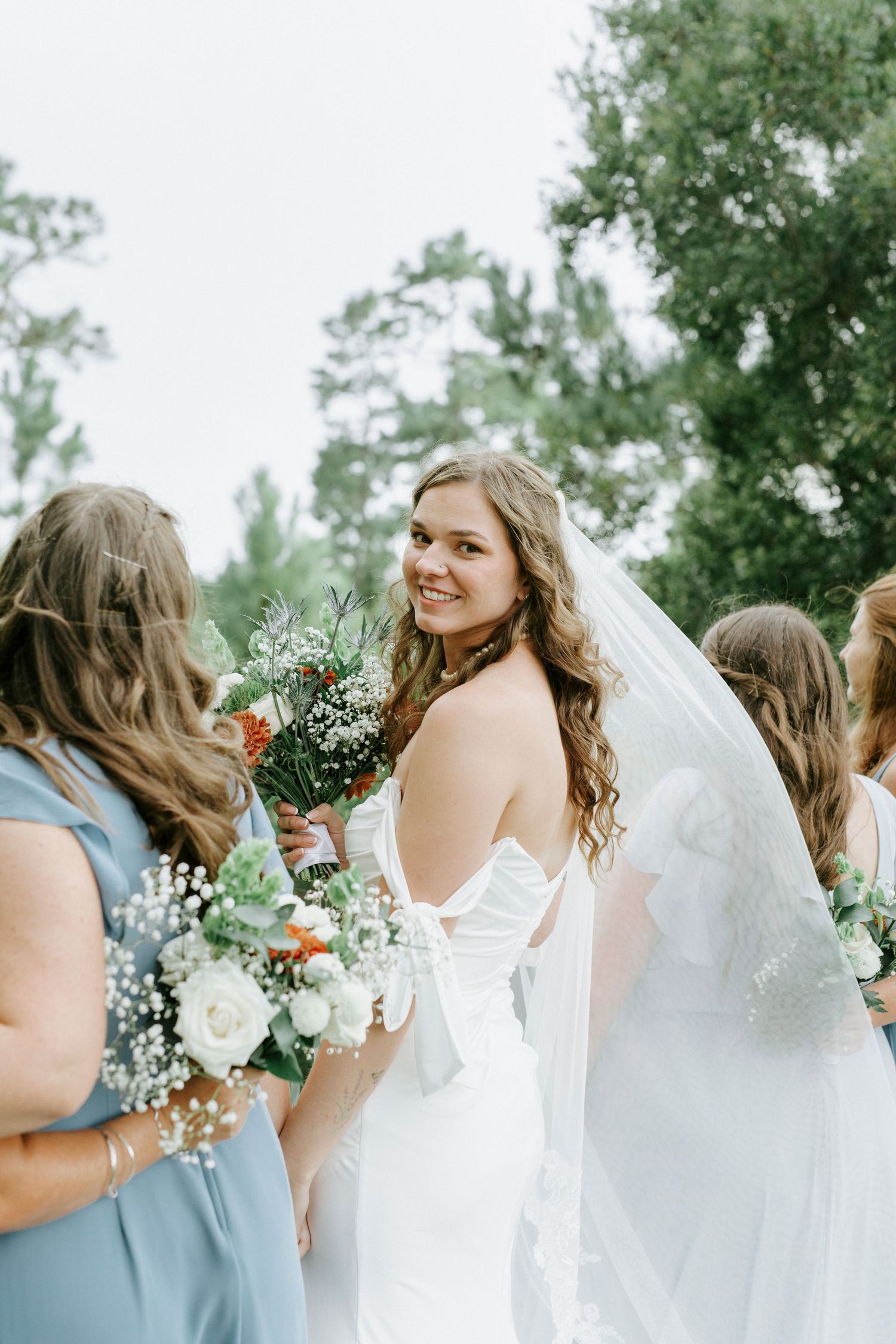Wedding party in blue and white dresses share joyful candid moments while holding floral bouquets outdoors.