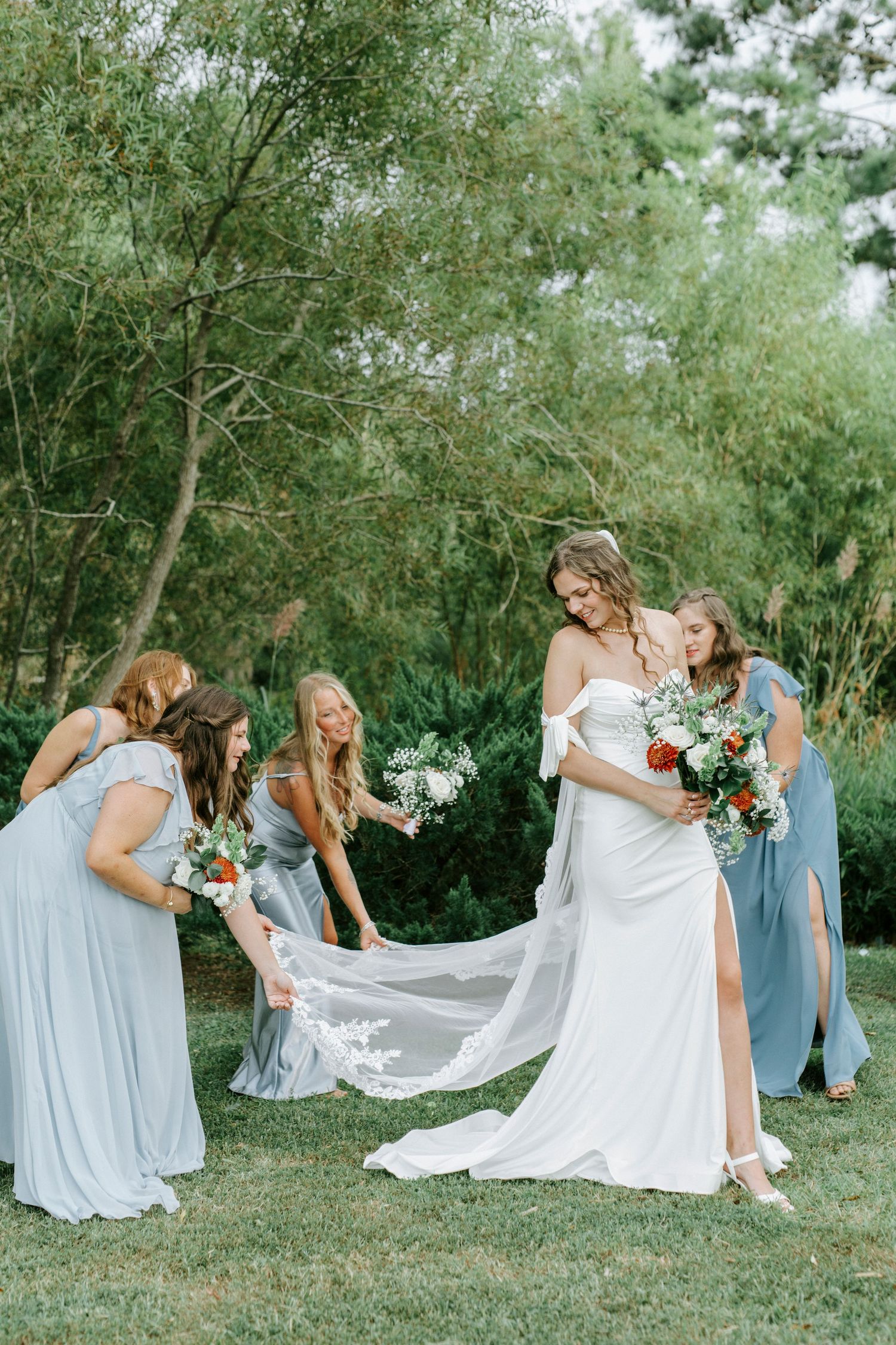 Bridesmaids in light blue dresses help arrange the bride's flowing white wedding gown train in an outdoor garden setting.