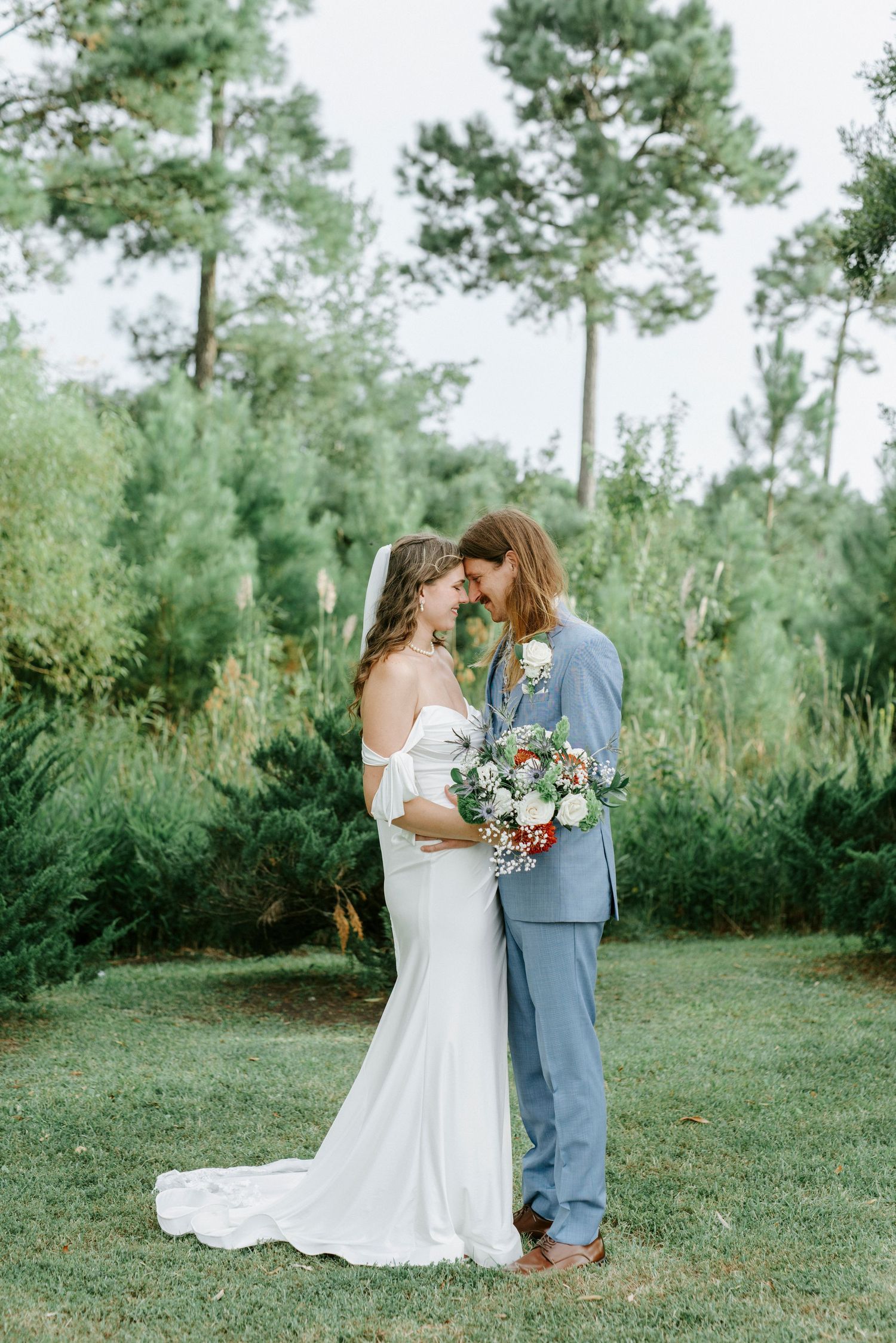 A romantic outdoor wedding scene with a couple embracing in a wooded setting with pine trees in the background.