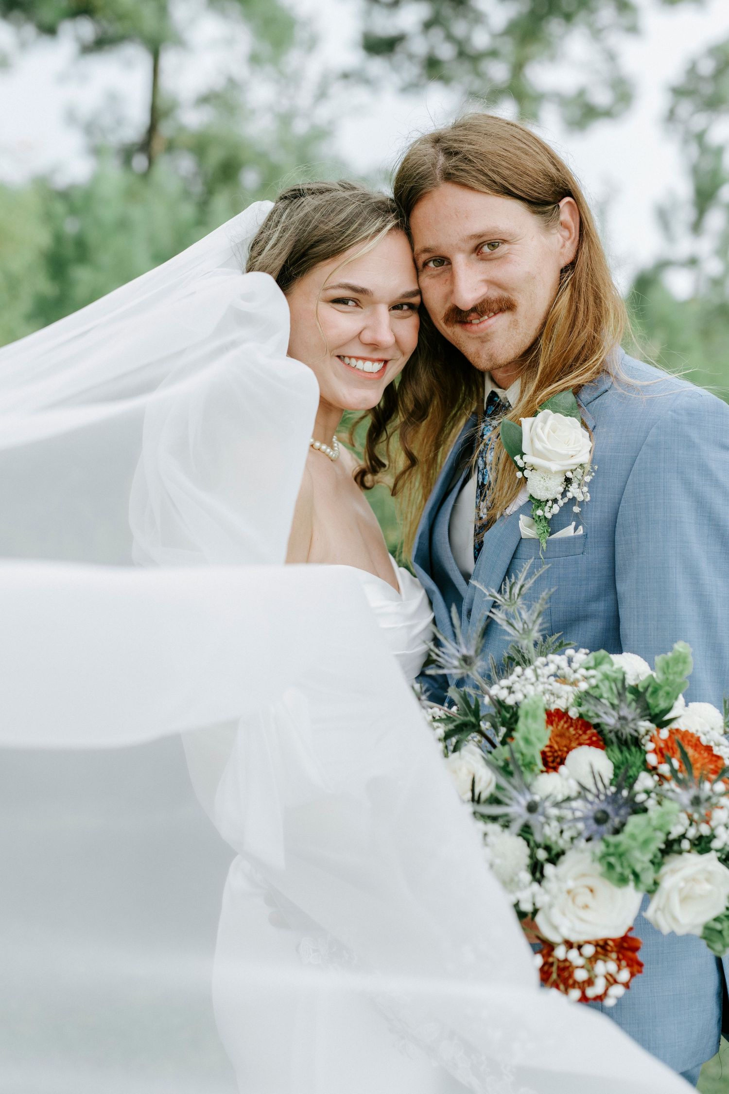 A joyful wedding couple poses together outdoors with a flowing white veil and beautiful floral bouquet.