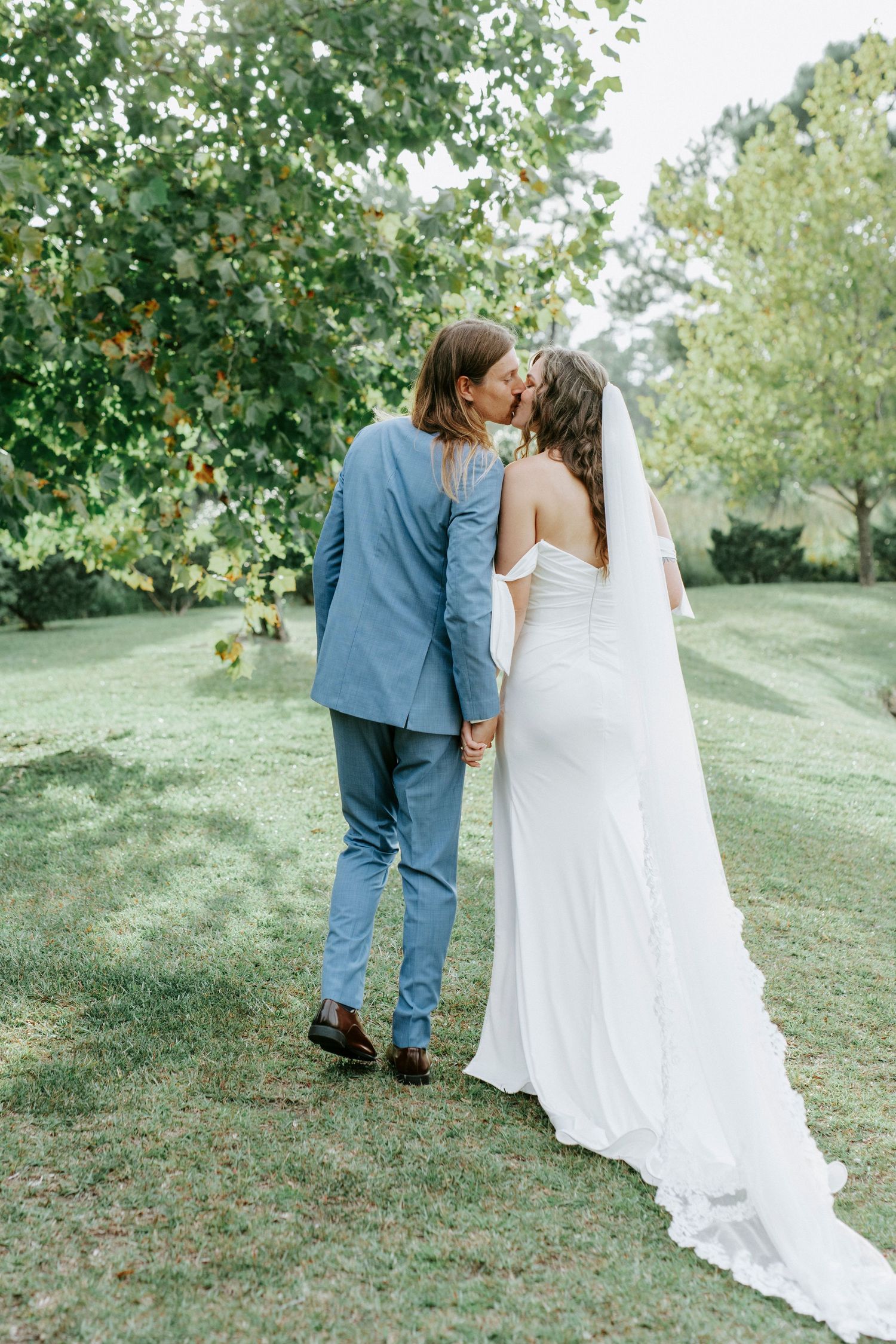 A couple in wedding attire shares an intimate moment on a grassy hillside surrounded by green trees.