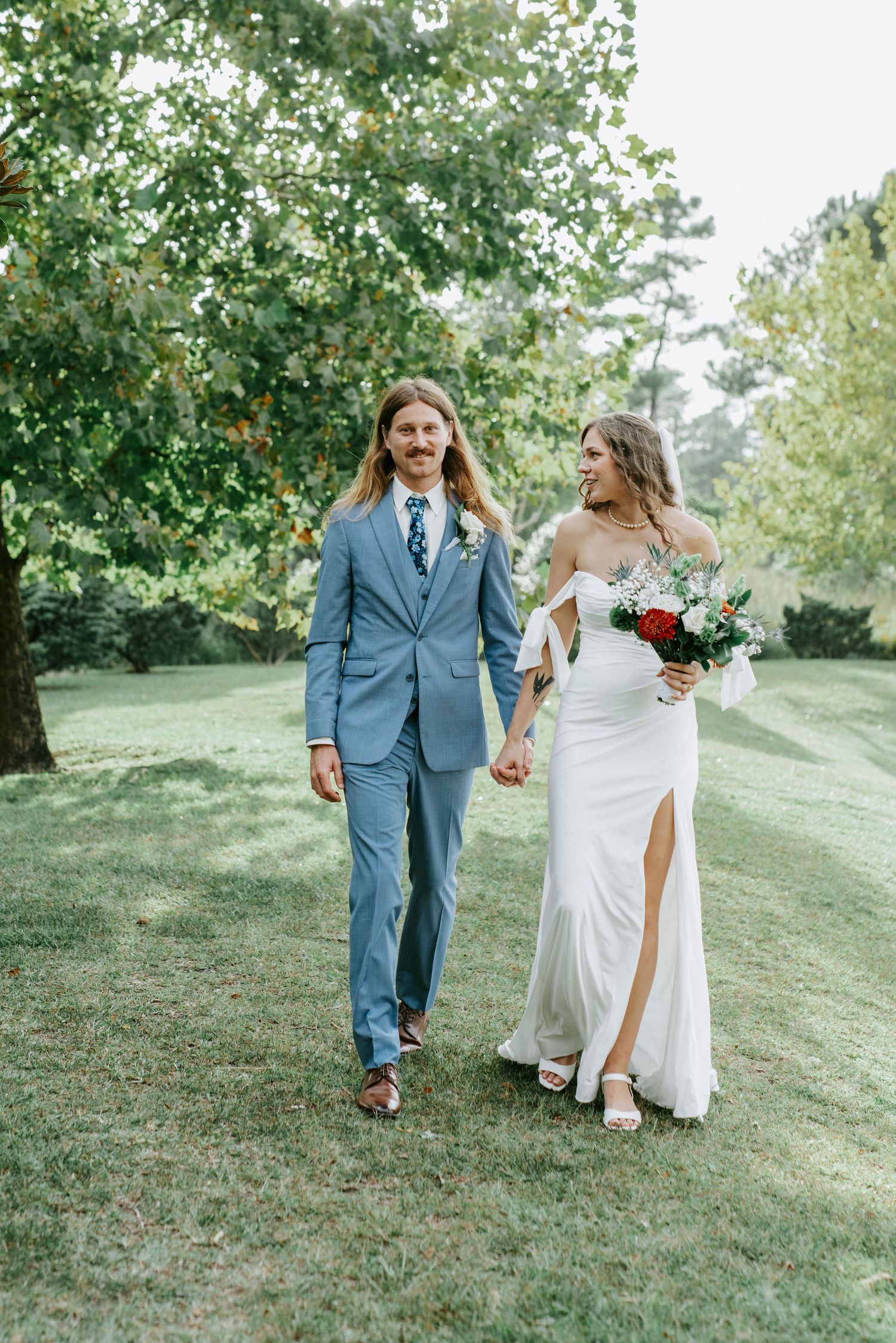 A couple in wedding attire walk hand in hand on a grassy hillside surrounded by green trees.