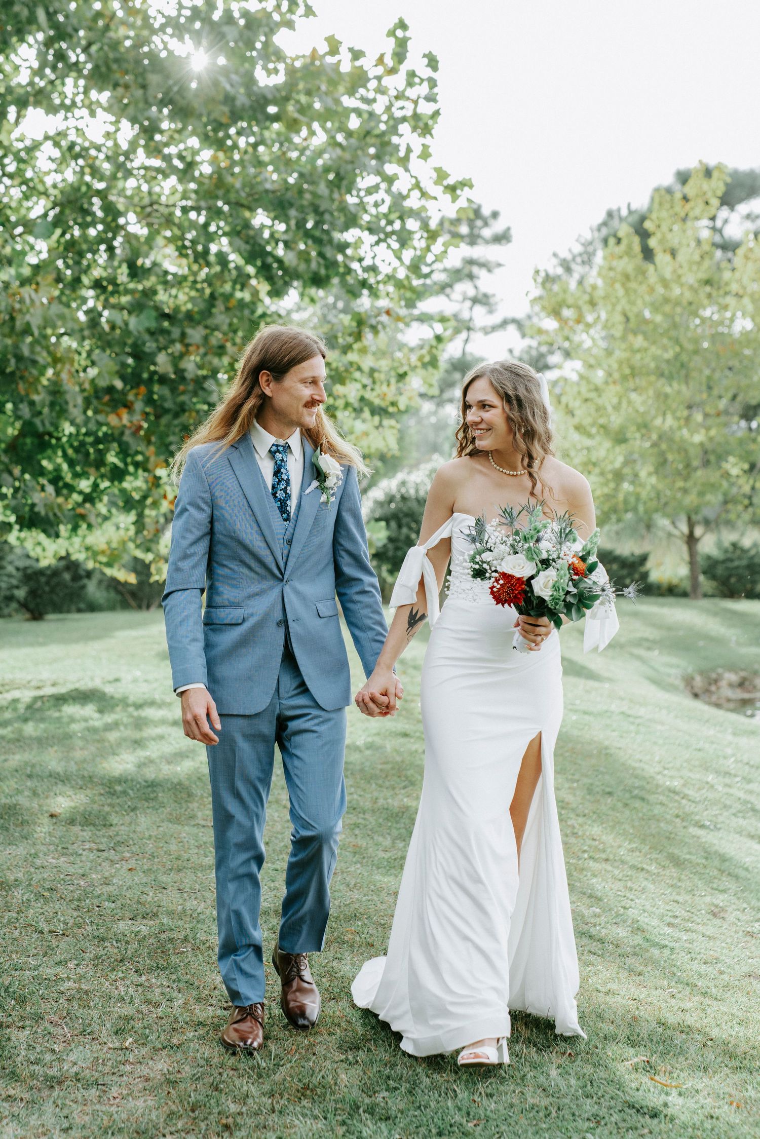A bride and groom walk together on a grassy hillside in coordinating blue suit and white wedding dress.