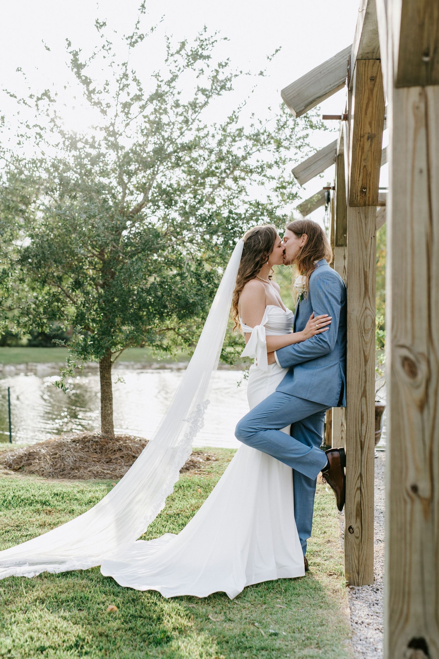 A romantic wedding couple embraces in a garden setting with flowing white dress and wooden posts framing the scene.
