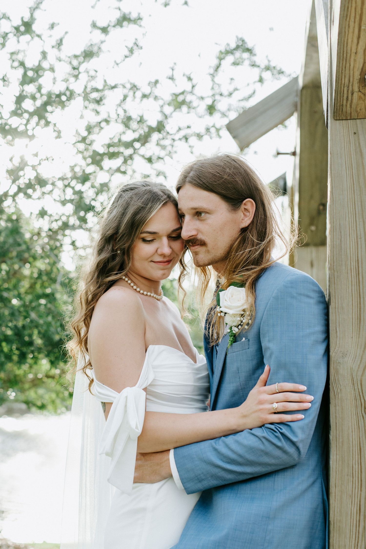 A romantic wedding couple embraces outdoors in soft natural lighting, with the bride in white and groom in light blue suit.