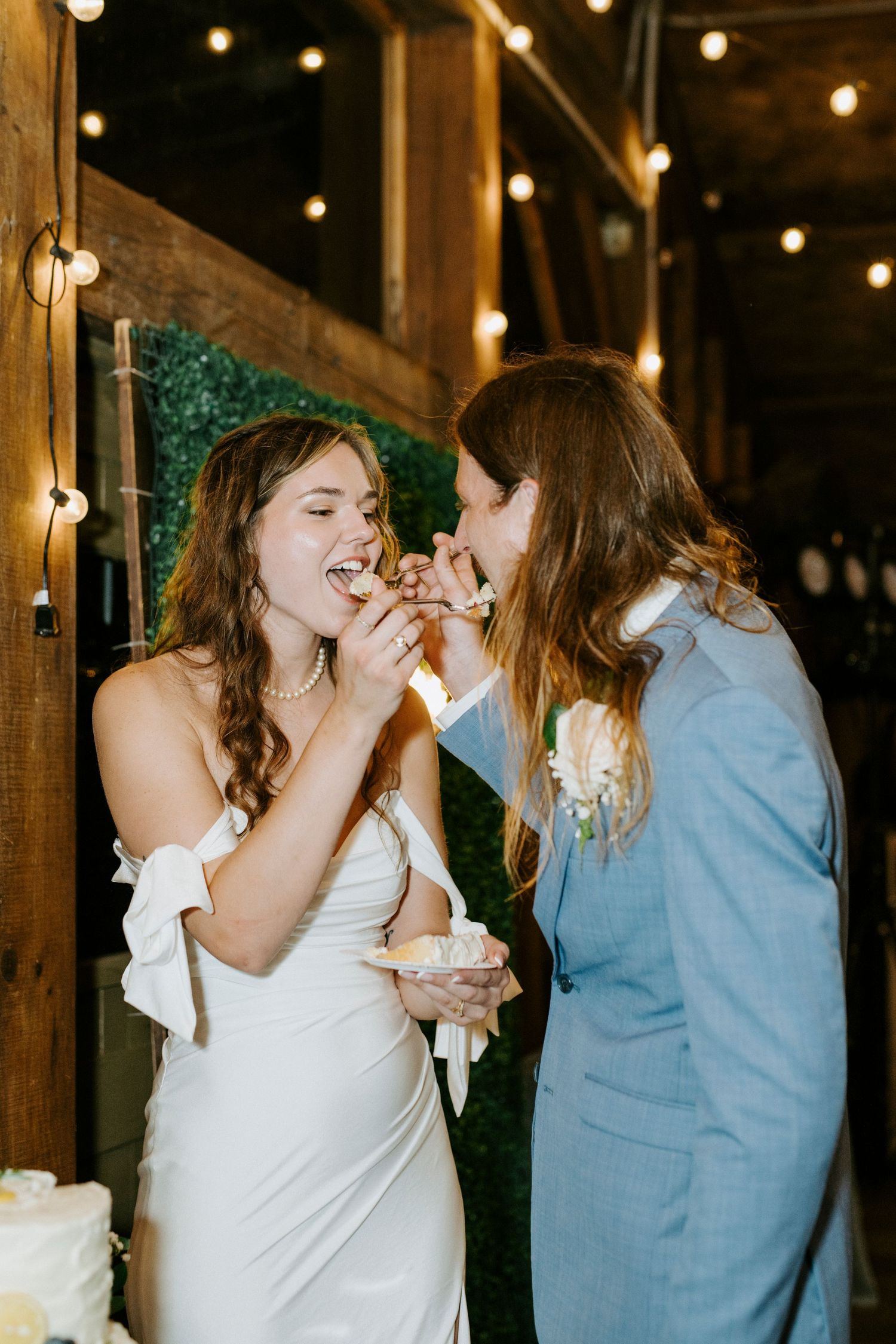 Two people in wedding attire share a joyful moment while eating cake together under string lights at a reception.