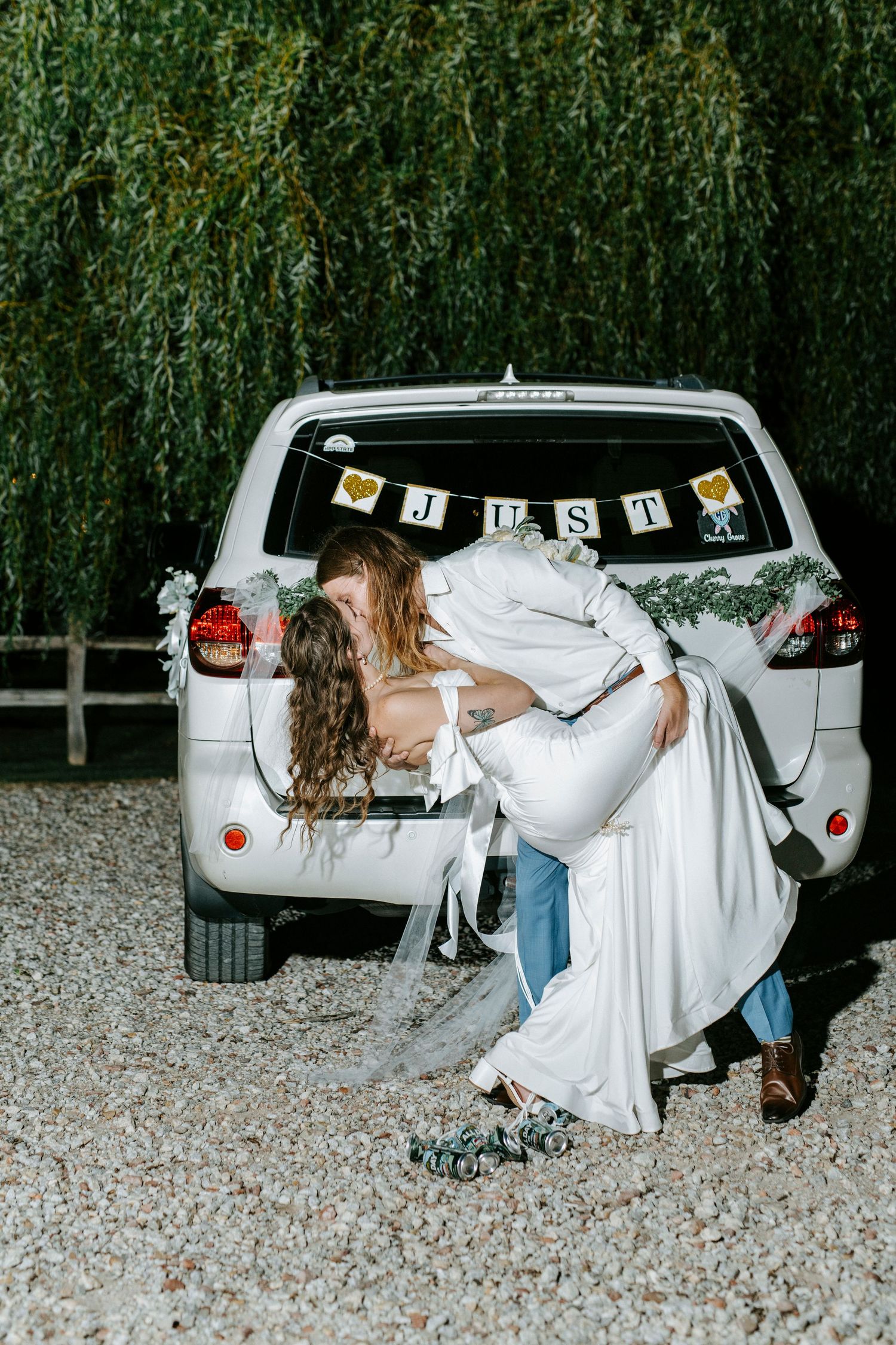 A white car decorated with wedding ribbons and 'Just Married' text parked in front of green foliage.