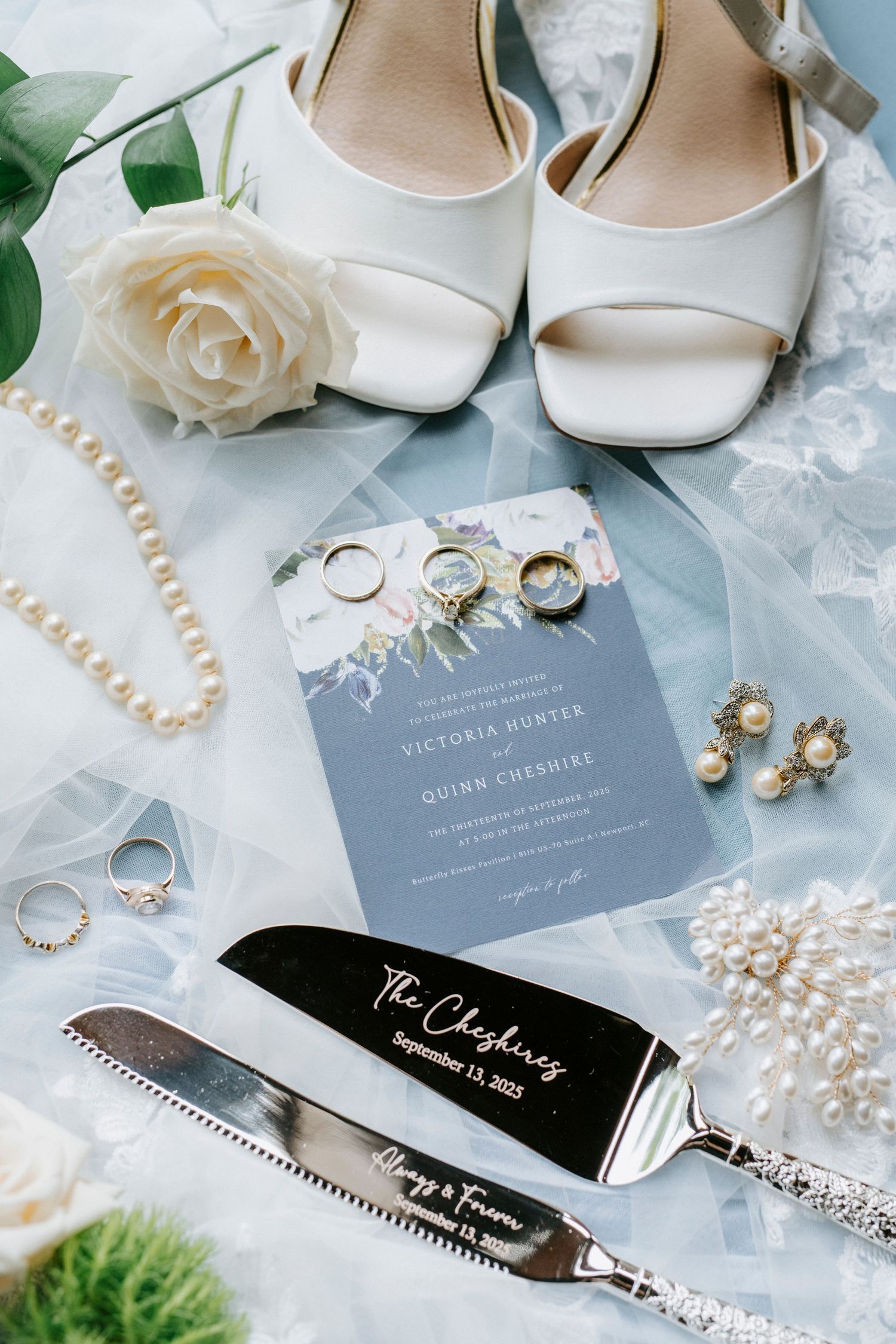 Wedding details flatlay with white bridal shoes, rings, invitation, pearl necklace, and cake serving set on lace background.
