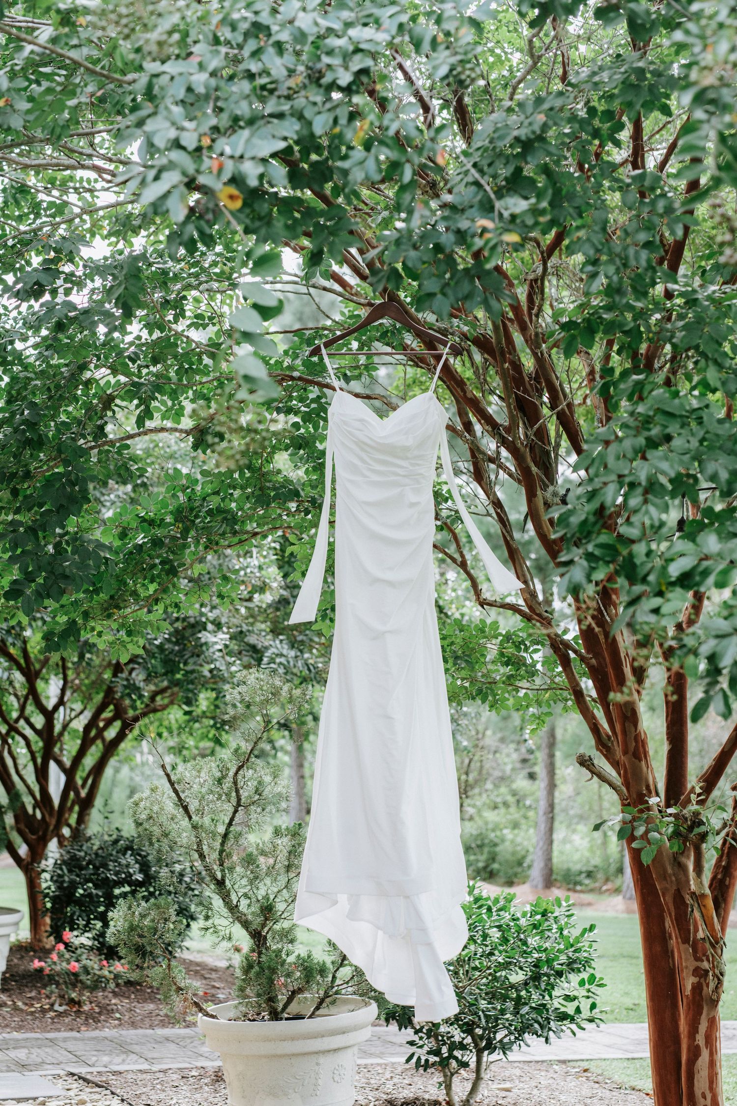 A white wedding dress hangs elegantly from a tree branch in a garden setting.