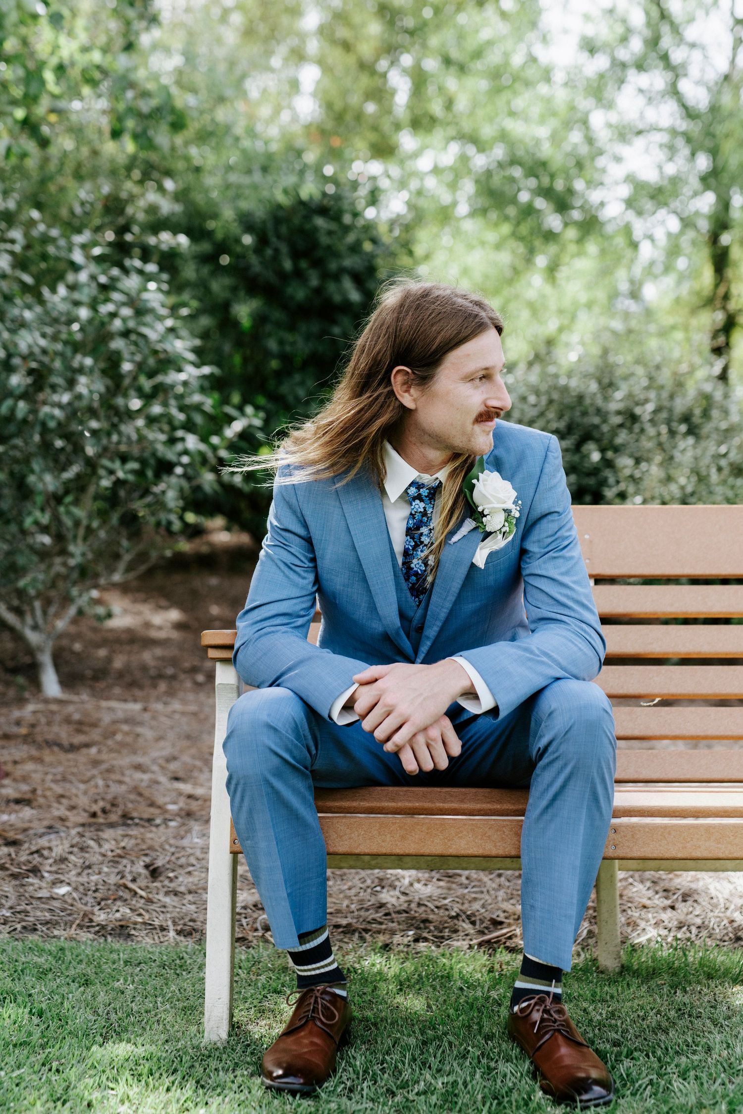 Person in blue suit sits contemplatively on wooden bench in garden setting.