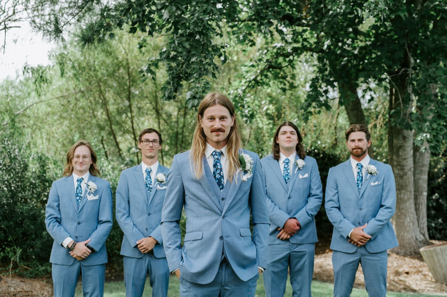 Groomsmen in light blue suits share a candid laughing moment together in an outdoor garden setting.